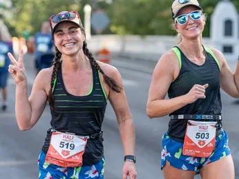 Two female runners smiling at the camera during a race, one giving a peace sign.