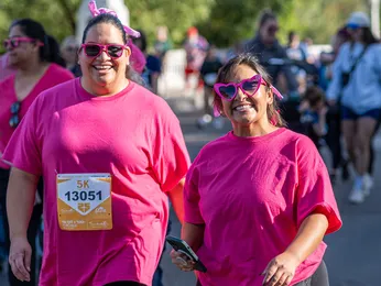 Two women in pink shirts and heart-shaped sunglasses smile at the camera while walking in a race.
