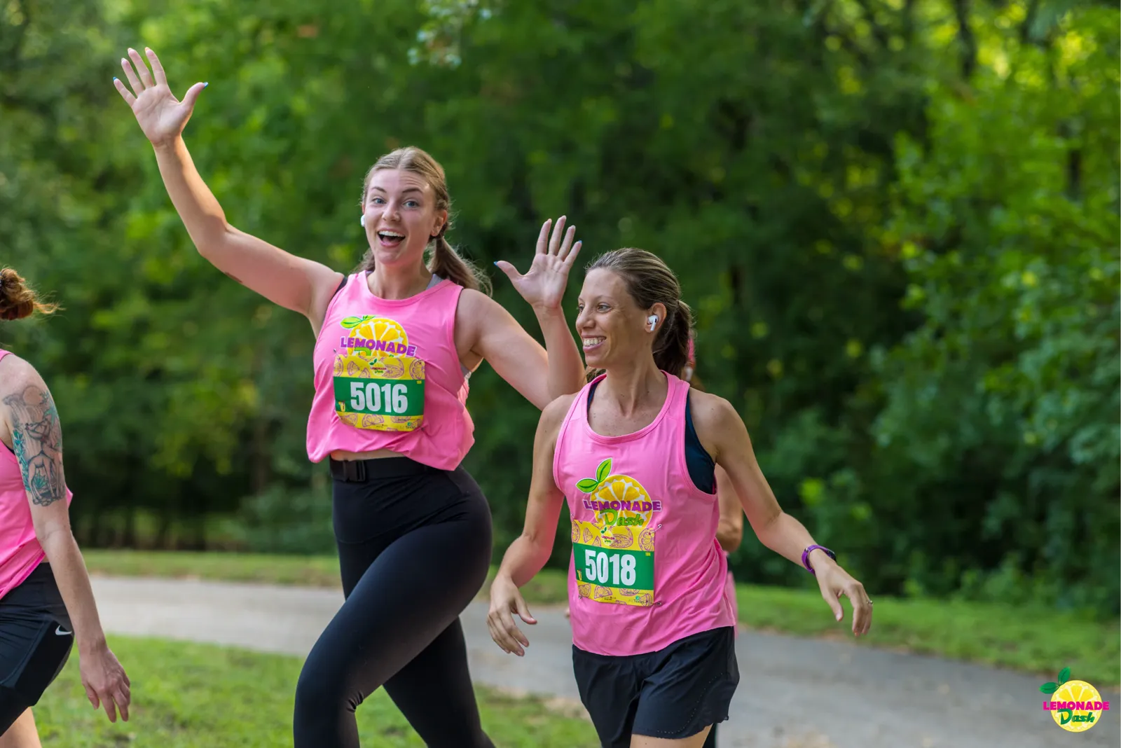 Two female runners in pink 'Lemonade Dash' shirts wave enthusiastically while running on a paved path with trees in the background.