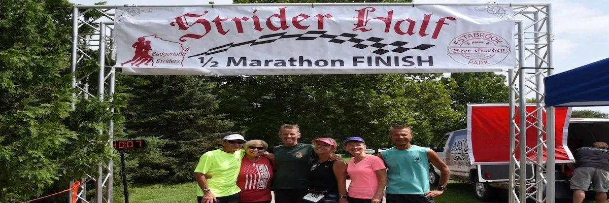 A group of six people smile for a photo under the Strider Half Marathon finish line banner.
