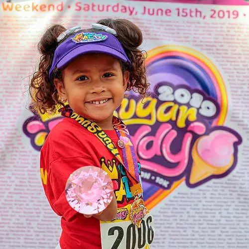 A young girl wearing a red shirt, a purple visor, and a race medal, holding a pink gem and smiling at the camera in front of a race banner.