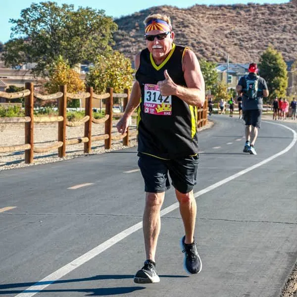 A male runner with bib number 3142 gives a thumbs up while running on a paved road during a half marathon.