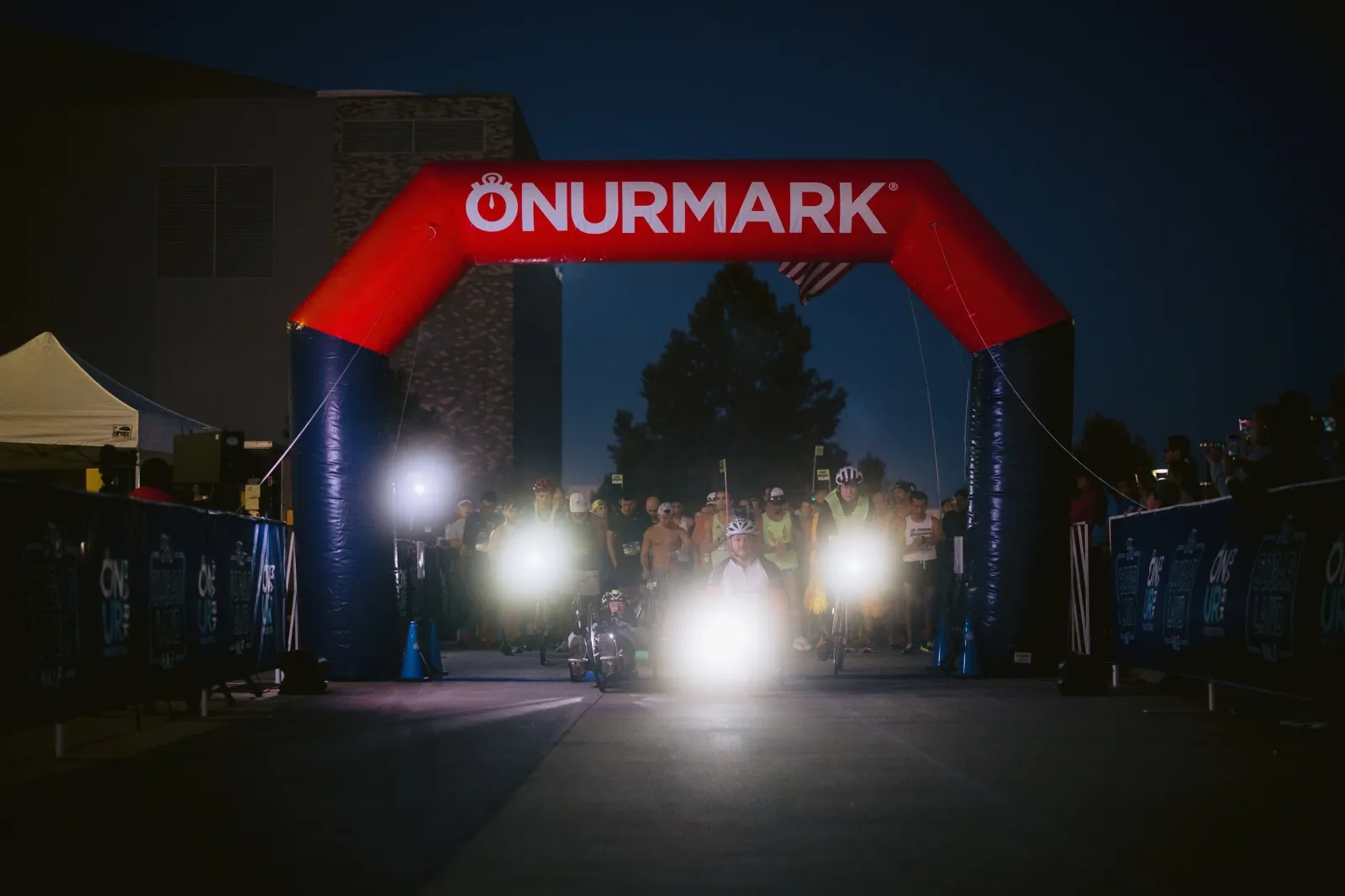 Runners gather at the start line under a large inflatable archway at night, with bright lights illuminating the front runners.