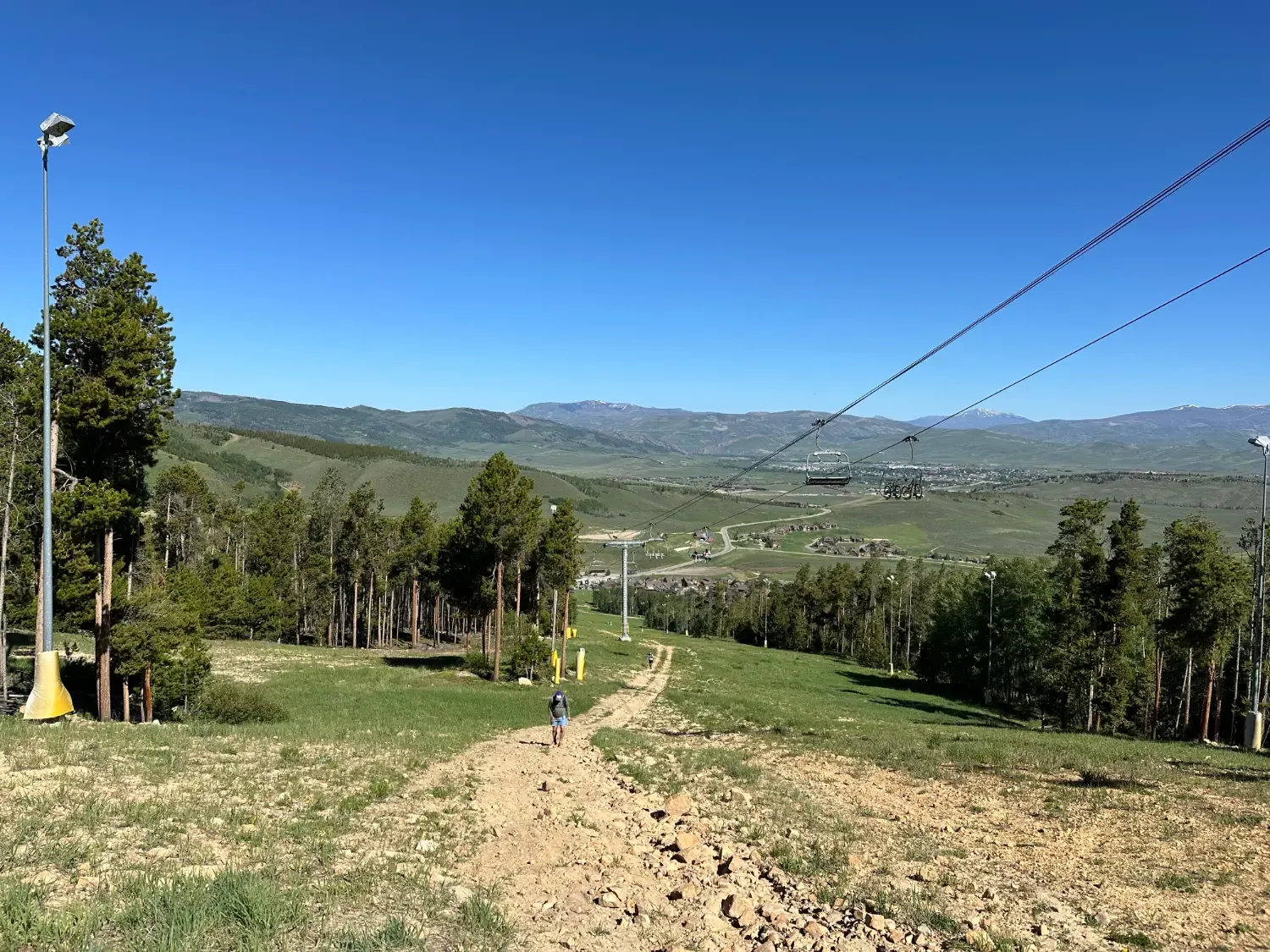 A lone runner walks up a dirt trail on a grassy mountainside with trees and a ski lift in the background.