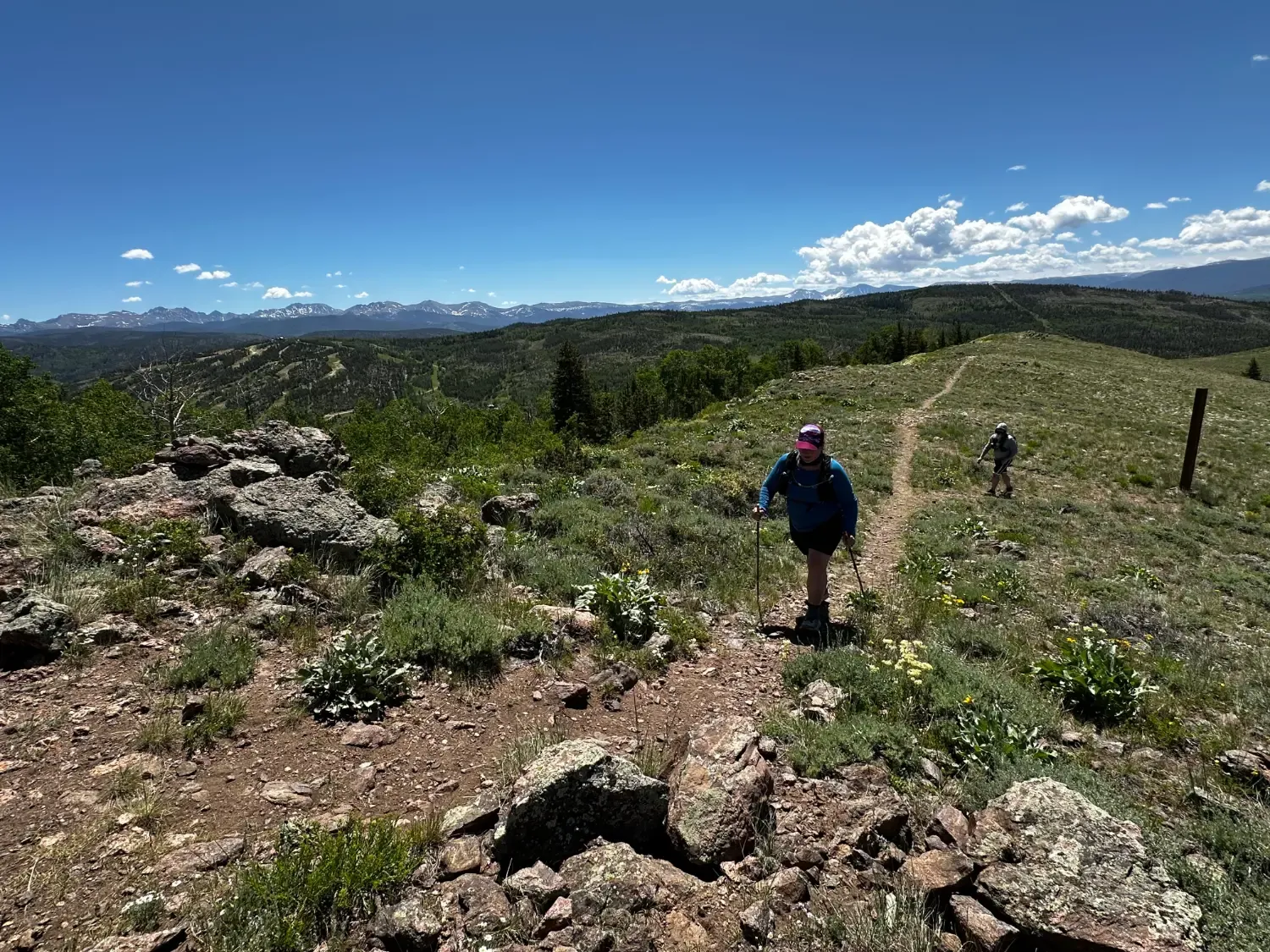 Two runners hiking on a winding trail through a mountainous landscape under a clear blue sky.