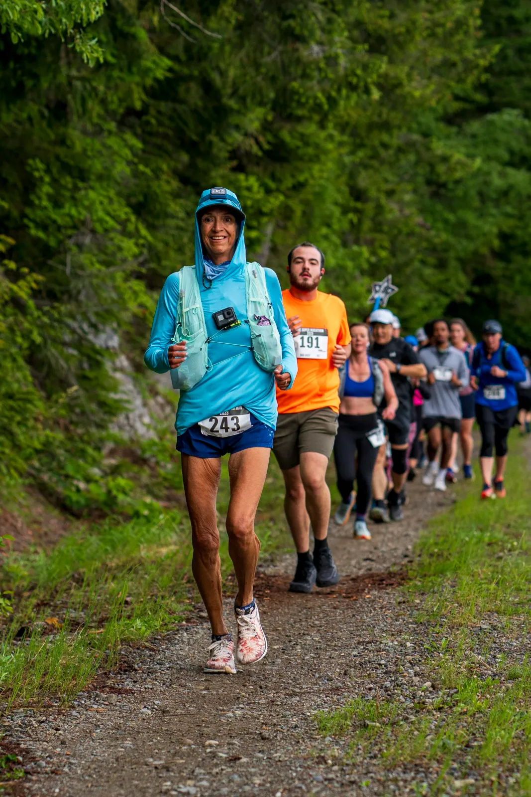 A female runner in a blue hoodie and shorts smiles as she runs on a gravel path, with other runners behind her on a tree-lined course.