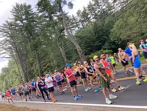 A large group of runners stand on a road, facing forward, ready to start a race.