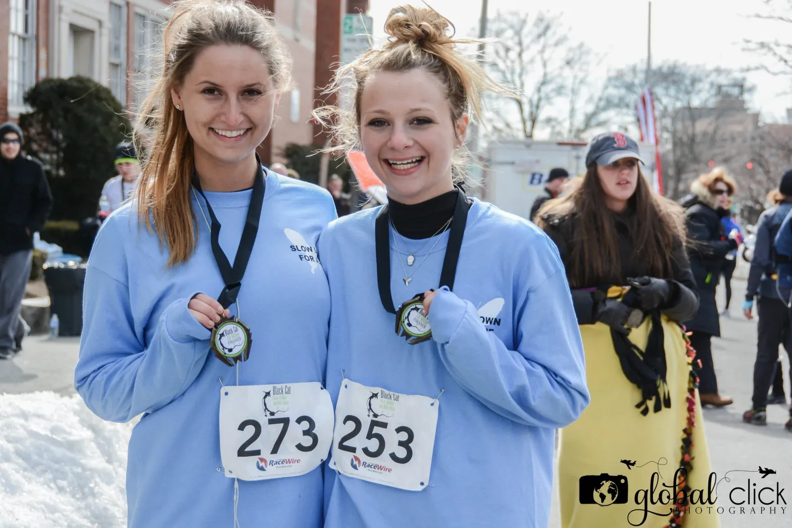 Two smiling women wearing blue long-sleeved shirts and race bibs, holding up their finisher medals.