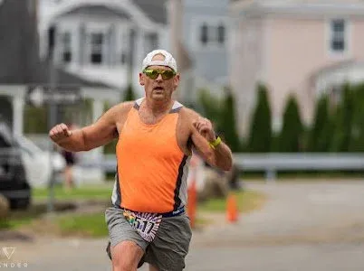 A male runner wearing an orange tank top and a white hat runs on a road during a race.