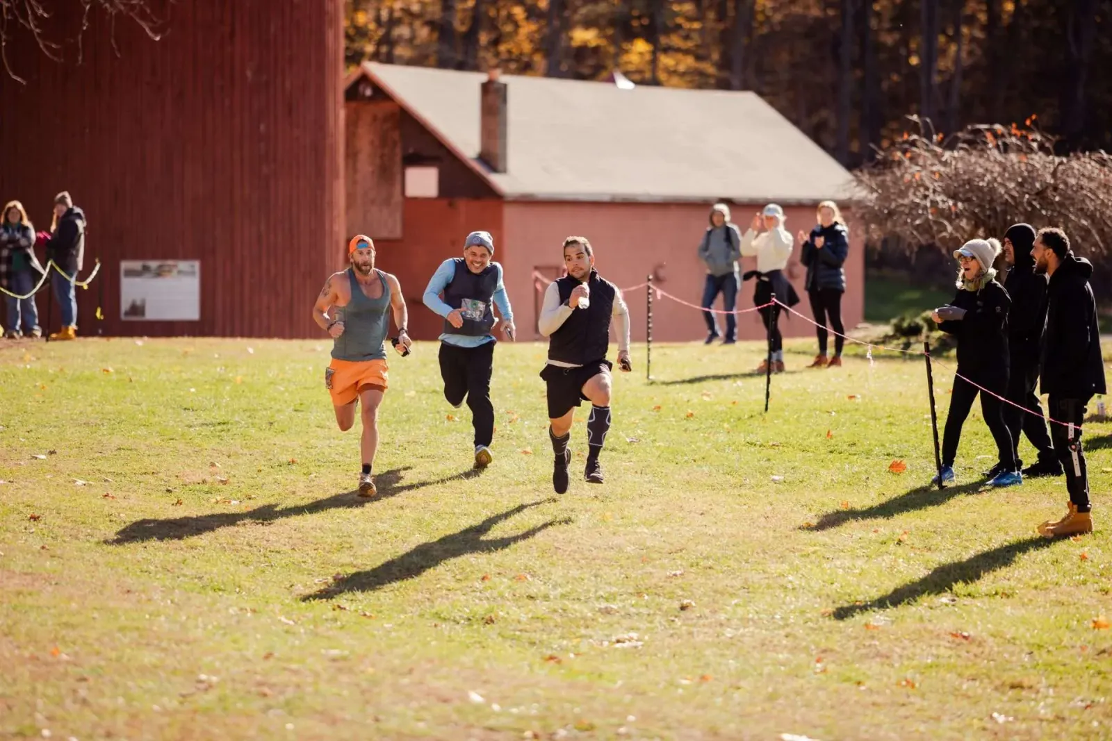 Three male runners are racing on a grassy field with buildings in the background.