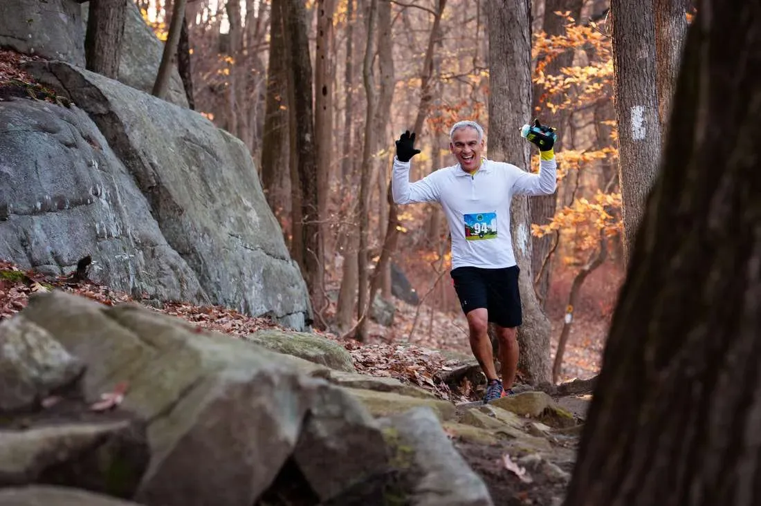 A smiling male runner with his arms raised in a celebratory gesture, running on a wooded trail during the Tarrywile Half Marathon.