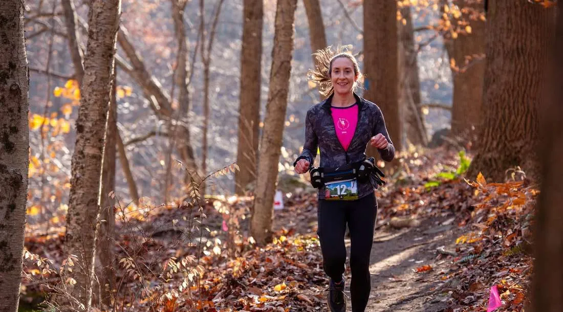 A female runner smiles as she runs on a wooded trail during a race.