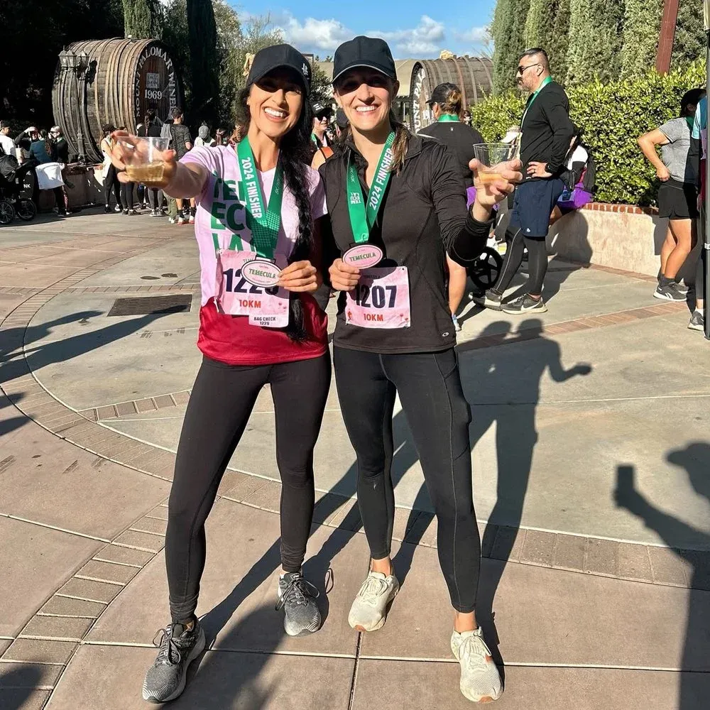 Two smiling women wearing race medals and holding drinks, celebrating after completing the Temecula Half Marathon, 10K, or 5K.