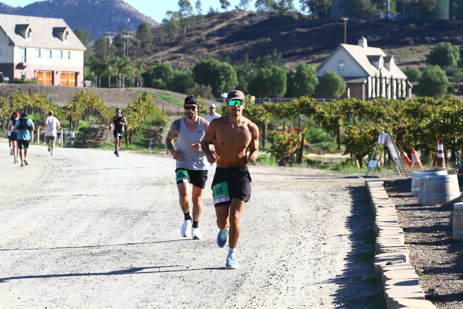 Two male runners are running on a dirt road with vineyards and buildings in the background.