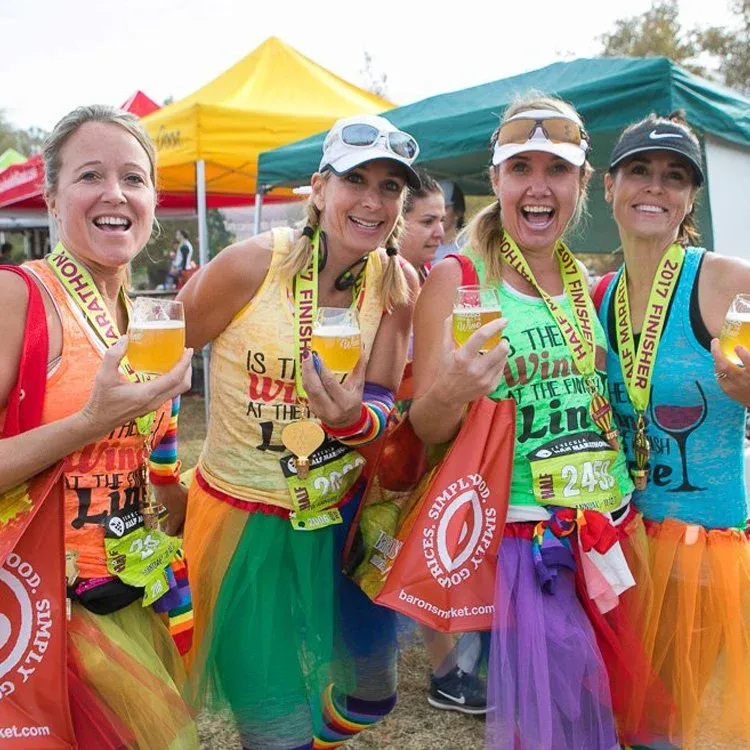 Four women in colorful tutus and race medals smile and hold up glasses of beer, celebrating after a race.