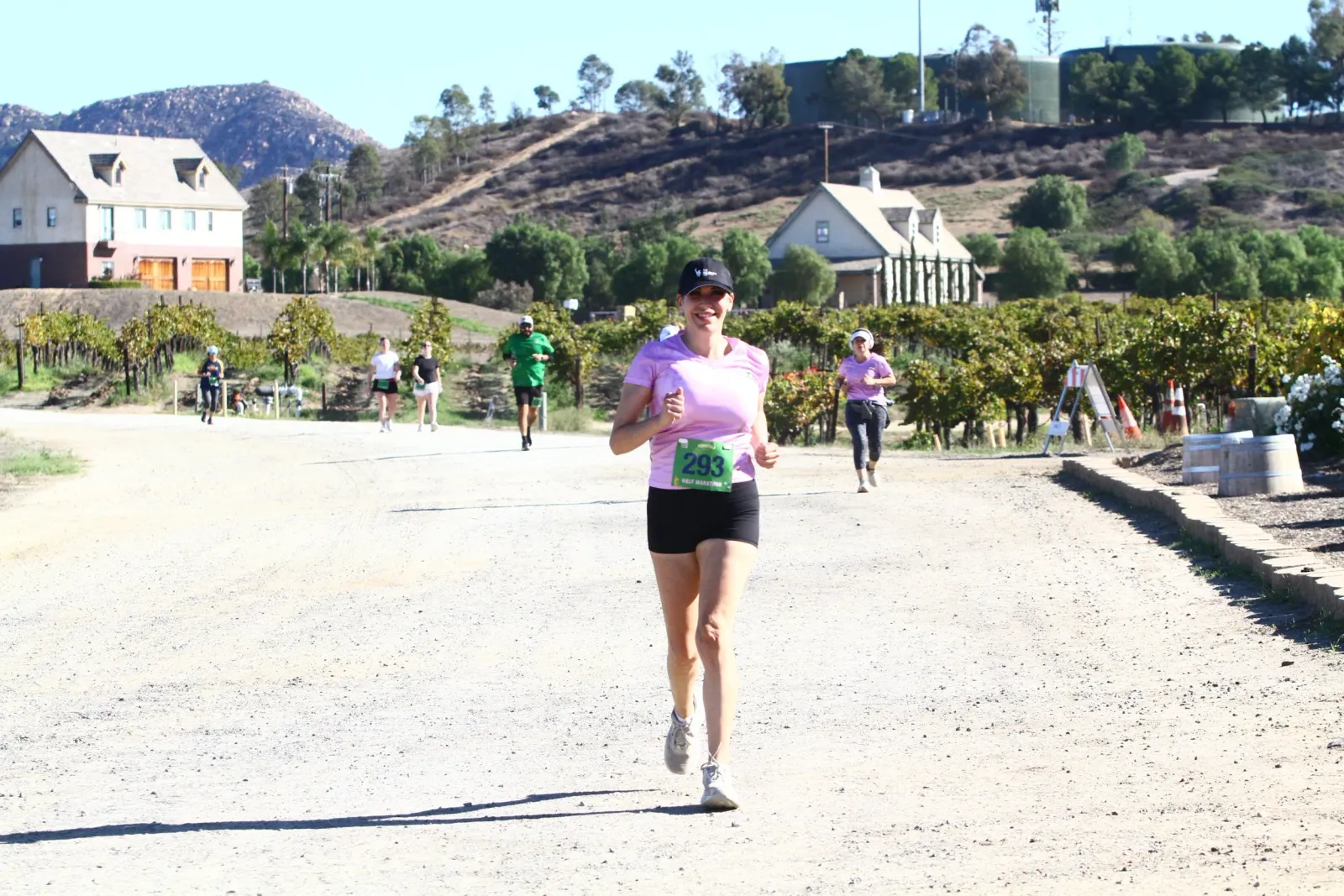 A female runner smiles as she runs towards the camera on a dirt road, with vineyards and houses in the background.