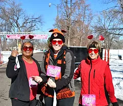 Three people wearing heart-shaped sunglasses and race bibs smile at the camera, with a race start line arch in the background.