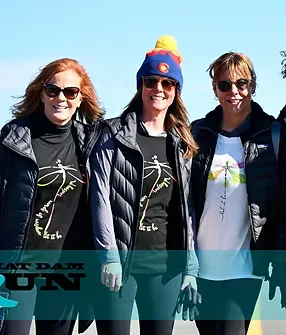 Three women wearing race t-shirts with a dragonfly logo and sunglasses, smiling at the camera, with a blue sky in the background.