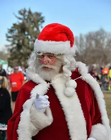 A person dressed as Santa Claus with a white beard and red hat, smiling and looking at the camera, with blurred runners in the background.