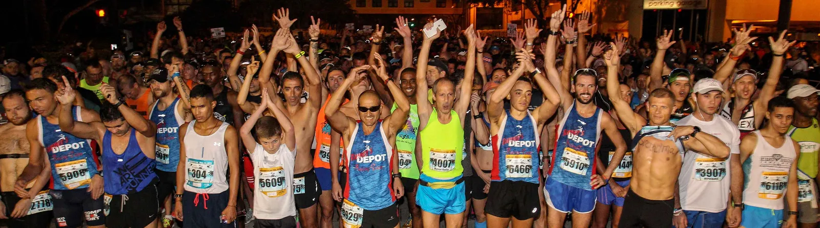 A large crowd of runners with their hands raised at the start of a night race.