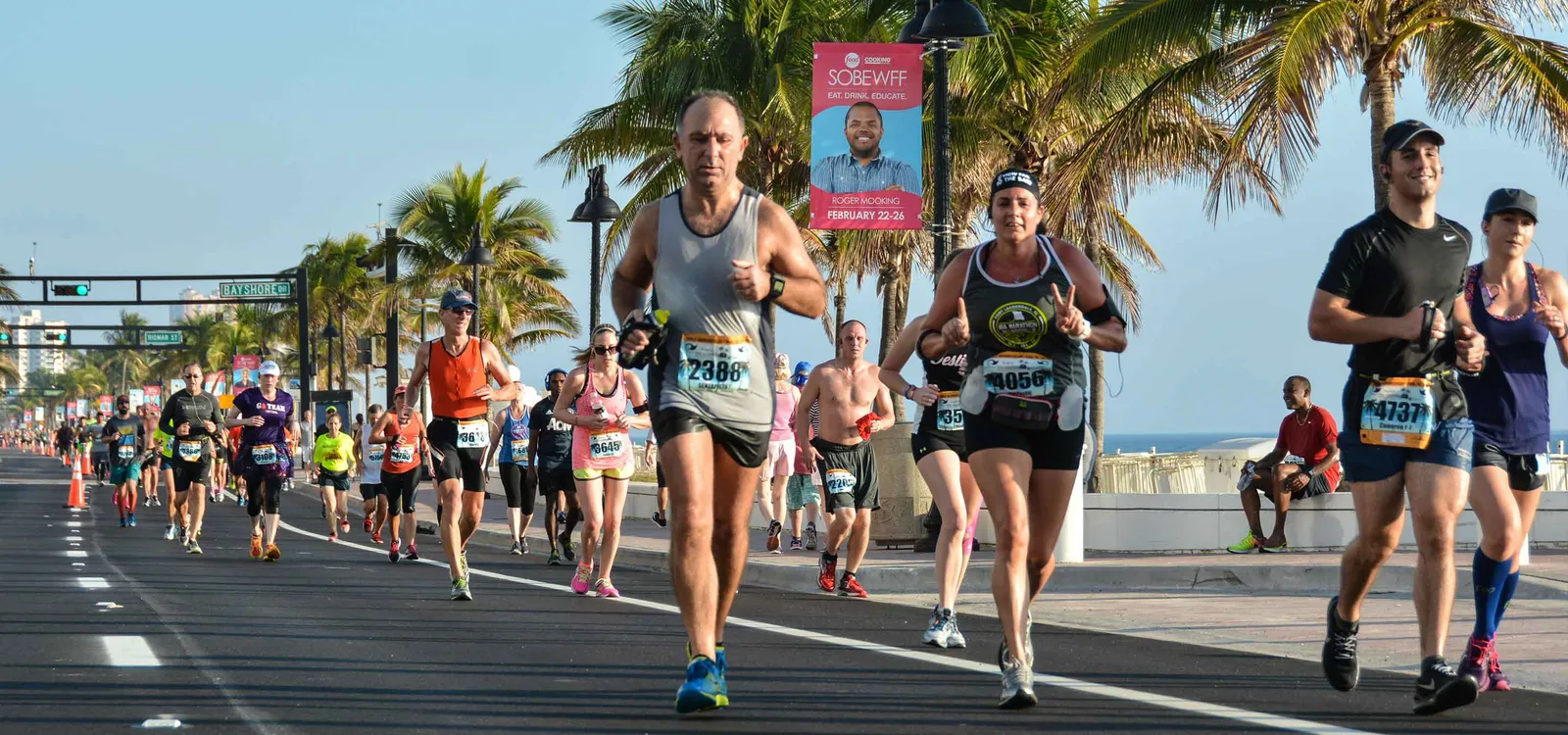 Runners participating in a race along a waterfront road with palm trees and buildings in the background.