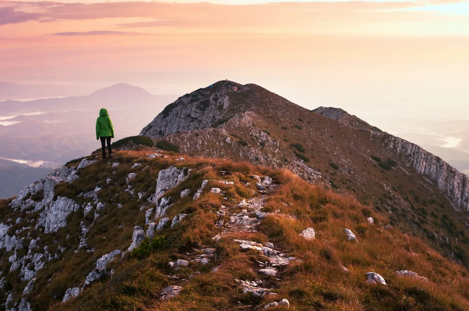 A person in a green jacket stands on a mountain ridge overlooking a misty valley at sunrise.
