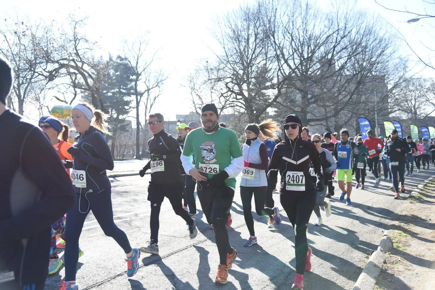 A group of runners participating in a road race, with trees and buildings in the background.