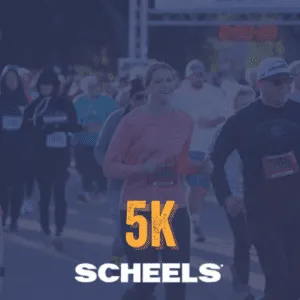 A crowd of runners, mostly smiling, are running towards the camera at the start of a 5K race. The word "5K" is prominently displayed in orange text, with "SCHEELS" below it in white.
