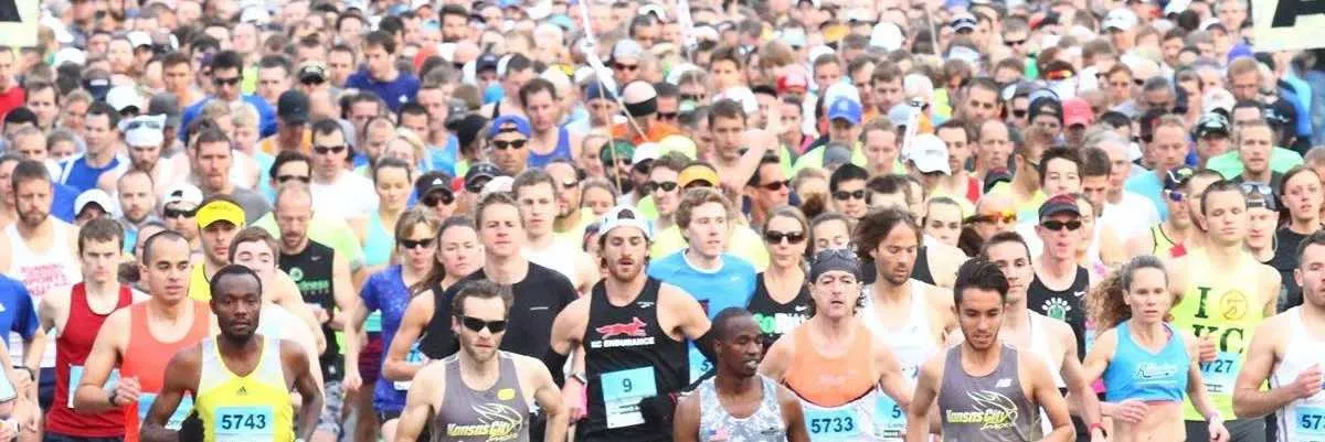 A large group of runners packed together at the start of a race, with many looking forward and some wearing race bibs.