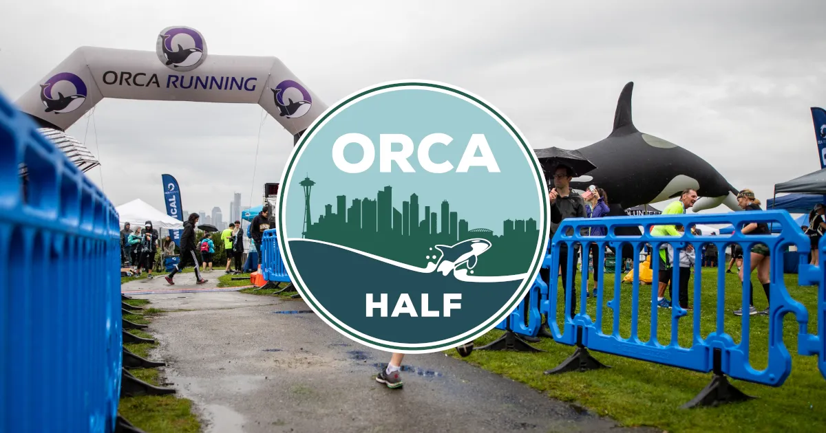 Runners at the start line of the Orca Half Marathon under an inflatable arch, with a large Orca mascot and the Seattle skyline in the background.