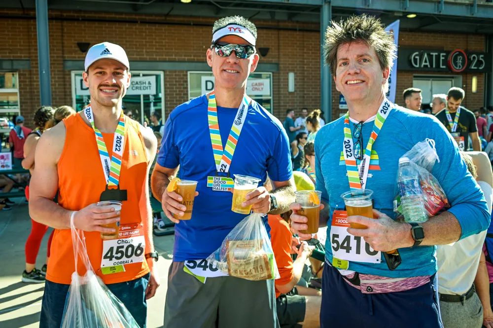 Three men wearing race medals and holding drinks, smiling after completing a half marathon.