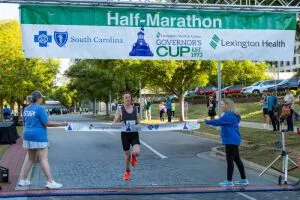 Runner crossing the finish line at the Half-Marathon Governor's Cup Road Race, with volunteers holding the banner.