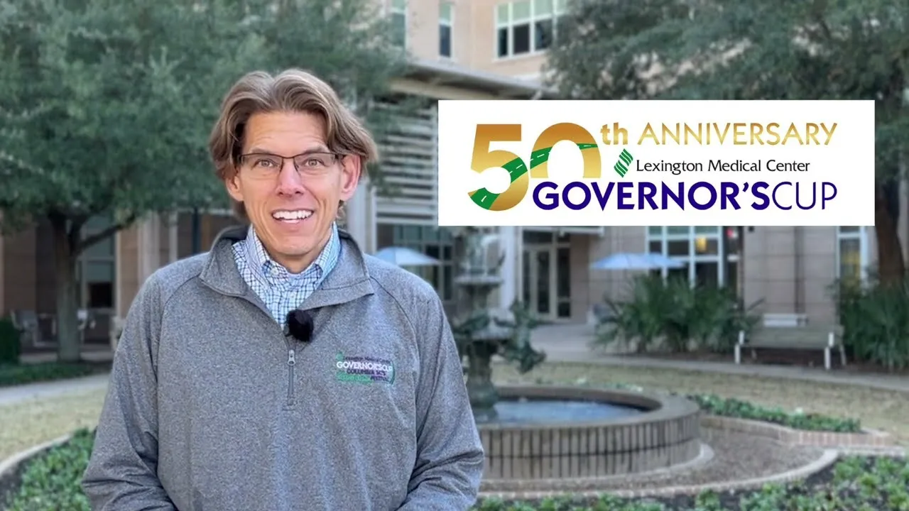 A man smiles at the camera, wearing a grey quarter-zip pullover with a race logo. Behind him is a building and a fountain, with a large overlay graphic for the 50th Anniversary Lexington Medical Center Governor's Cup.