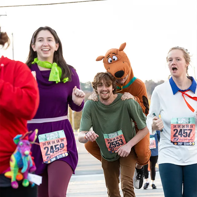 Runners in Scooby-Doo themed costumes participate in a Halloween race, with one person carrying another on their shoulders.