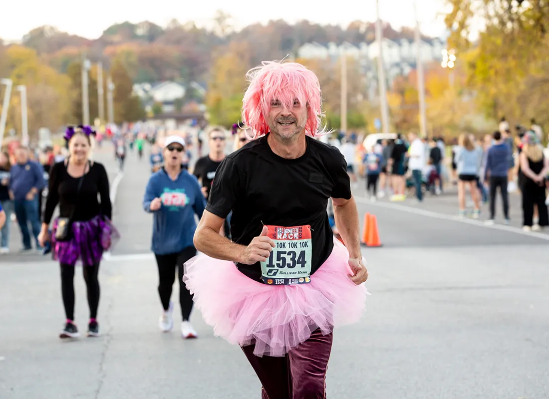 A runner wearing a pink wig and tutu gives a thumbs-up while running in a race, with other participants in the background.