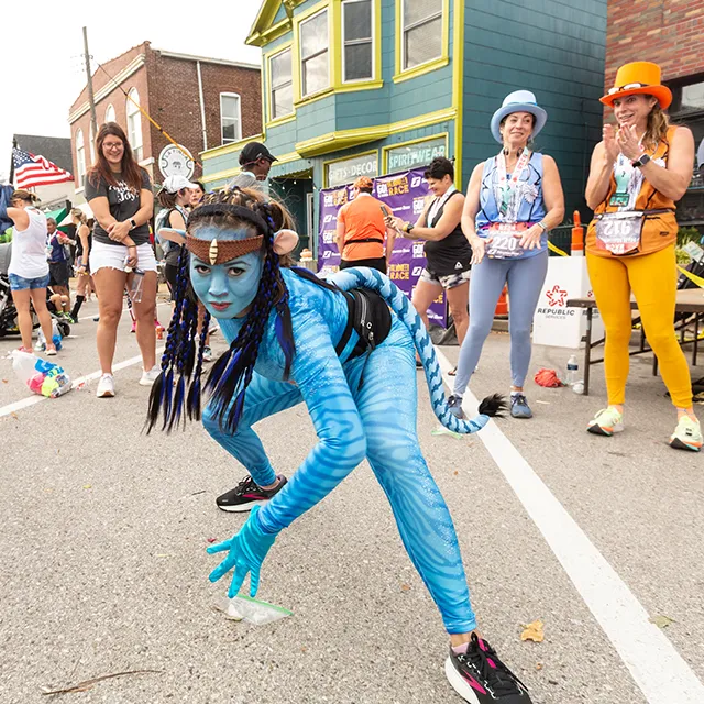A runner dressed as an Avatar character poses for a photo at the Great Go St. Louis Halloween Race, with other participants and spectators in the background.