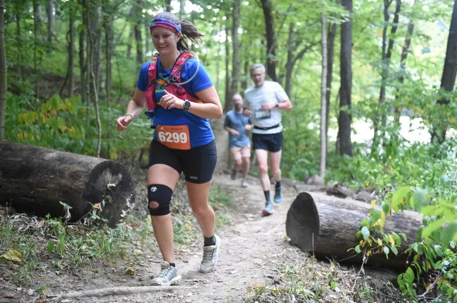 A female runner smiles as she runs on a wooded trail, wearing bib number 6299.