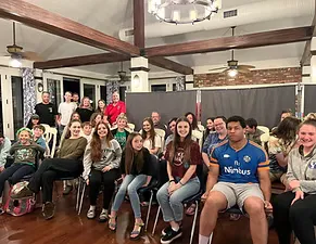 A diverse group of people, including children and adults, sitting and looking towards the camera in an indoor gathering space.