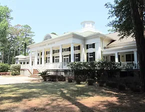 A large, white, colonial-style building with columns and a domed roof, set amidst trees and greenery.