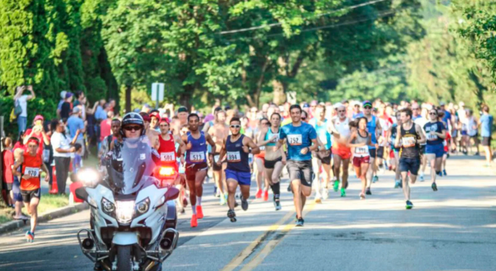 A large group of runners are participating in a race, led by a police motorcycle, with spectators visible on the side of the road.