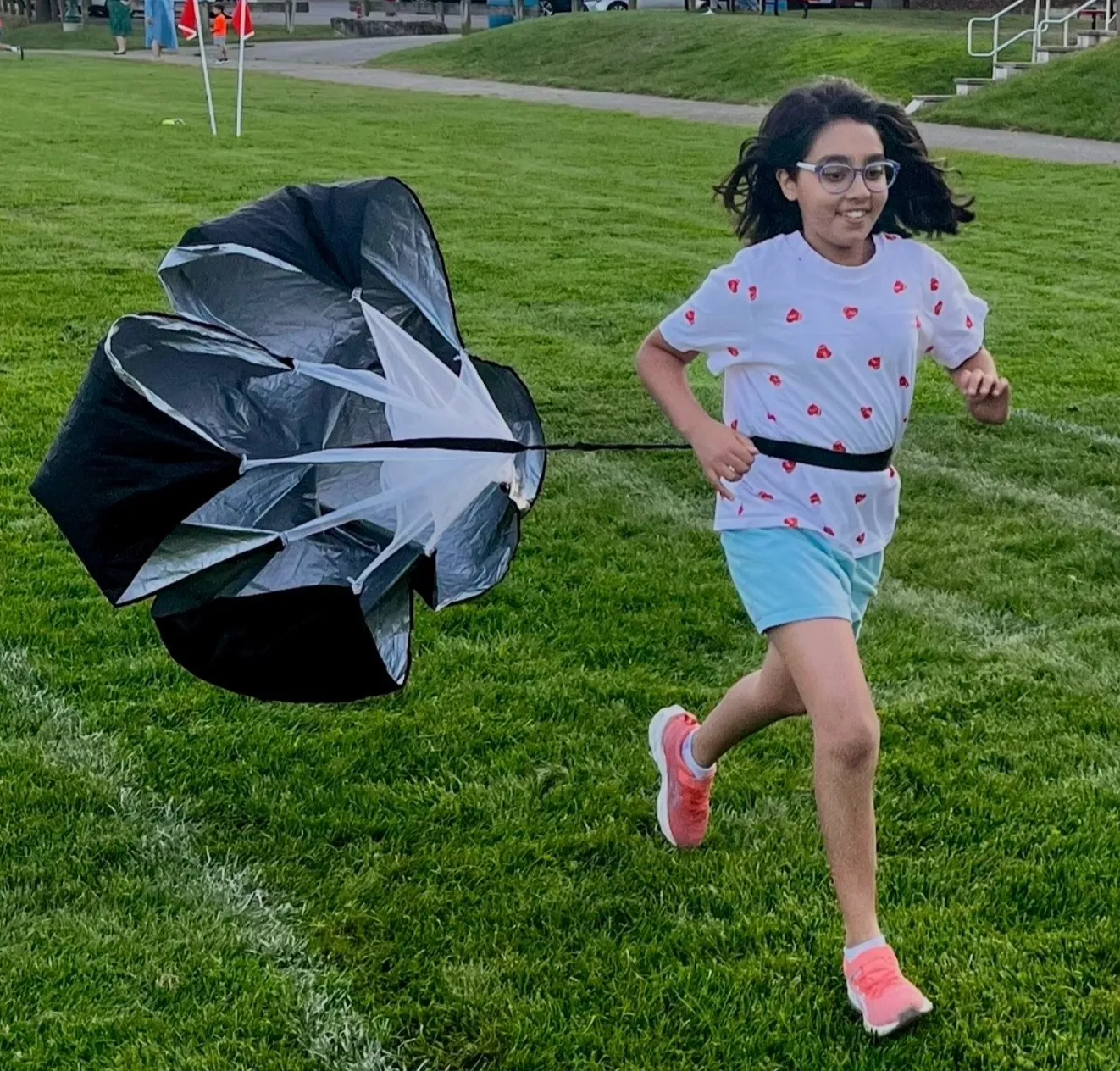 A young girl with glasses runs across a grassy field, pulling a black parachute behind her.
