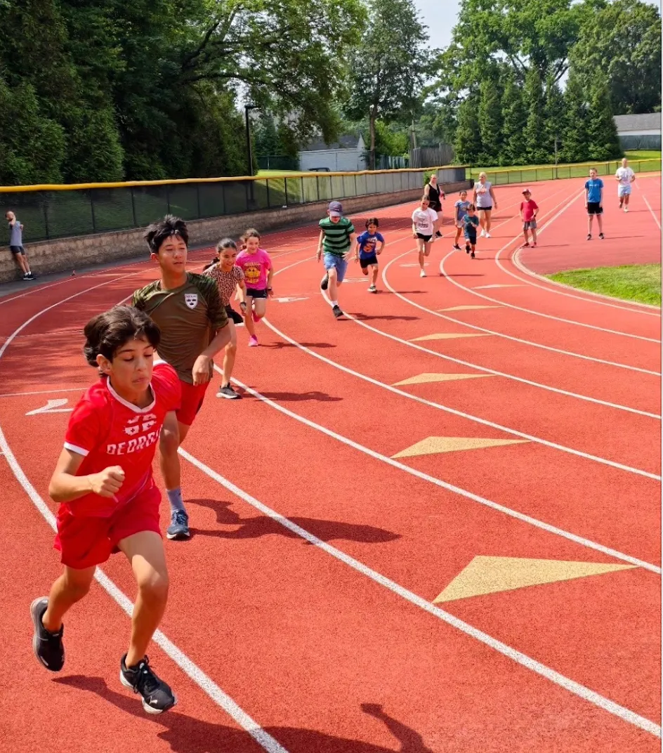 A group of young runners are racing on a red track, with one runner in the foreground wearing a red uniform.