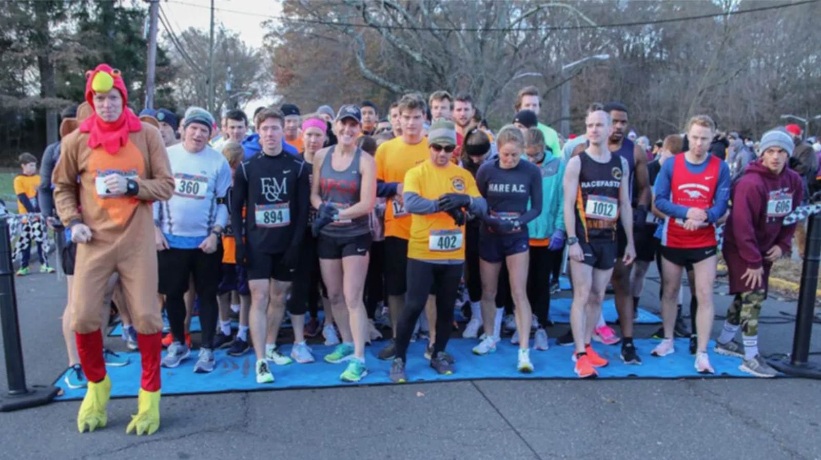 A group of runners at the start line of a race, with one runner in a chicken costume.