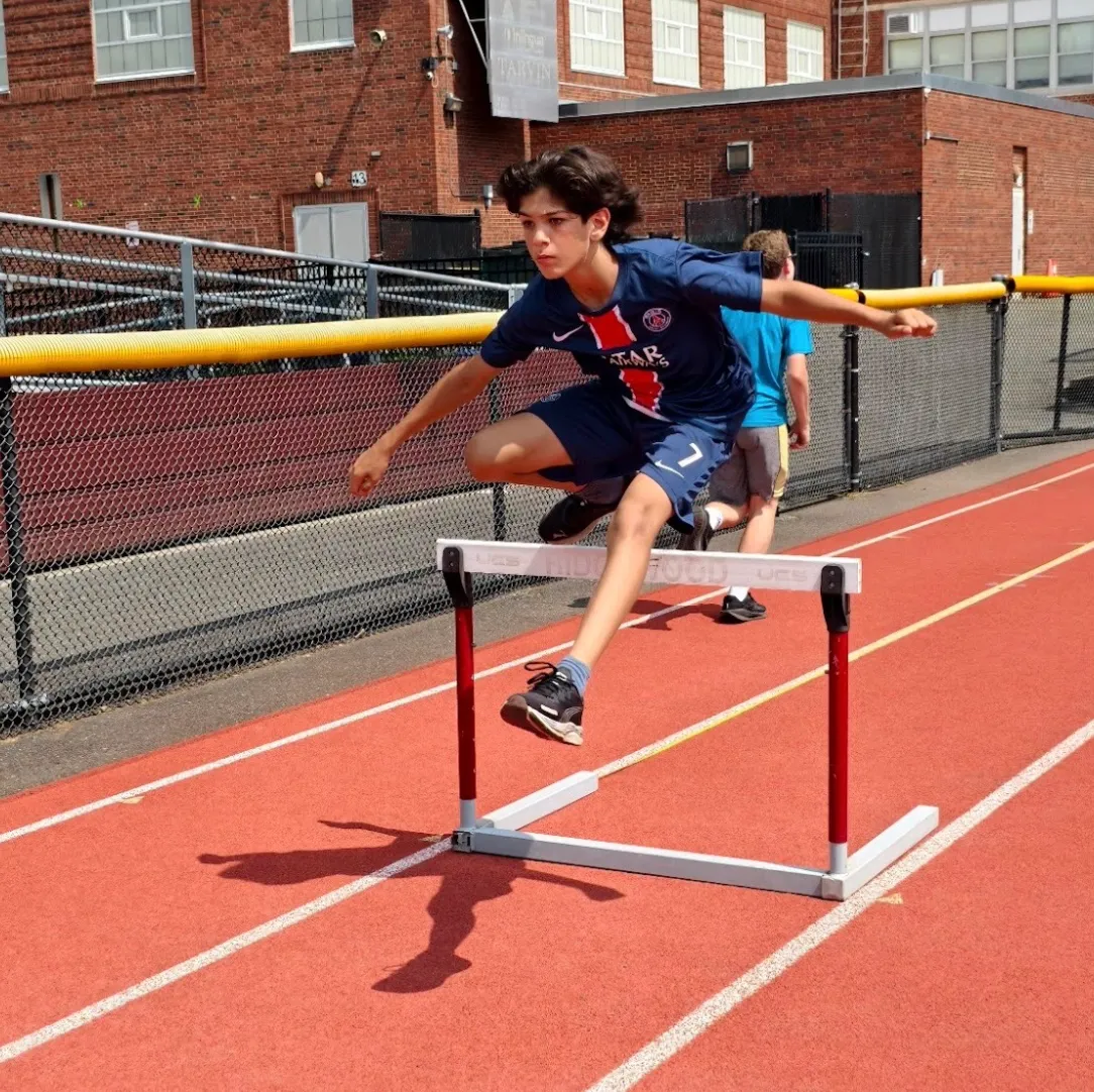 A young runner leaps over a hurdle on a track.