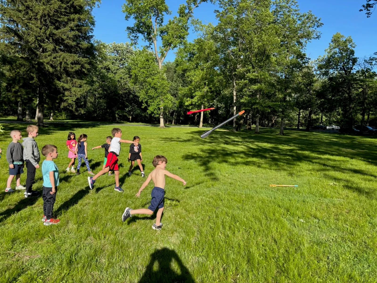 Children playing with javelins in a grassy park on a sunny day.