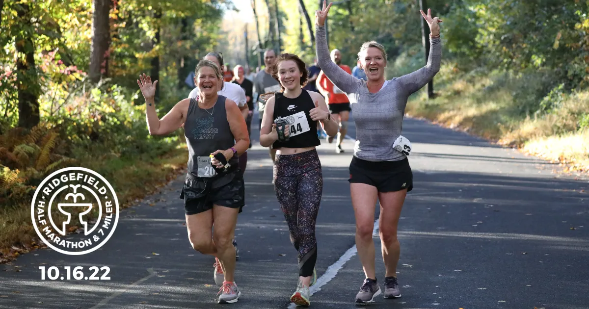 Three women running a race, two of them smiling and waving, with a third giving a thumbs up and a peace sign.