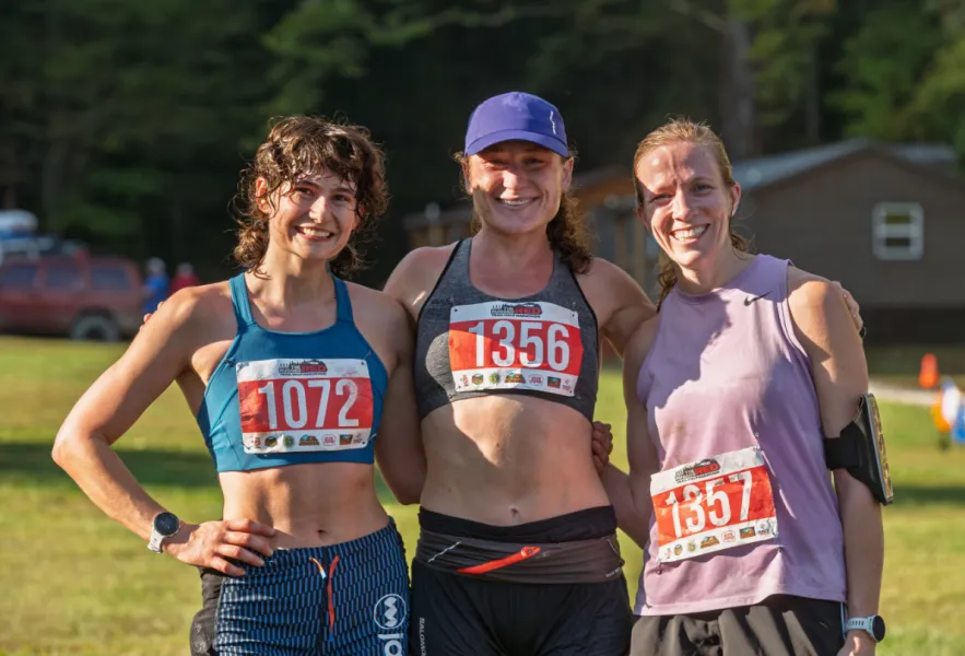 Three smiling female runners posing for a photo, wearing race bibs.
