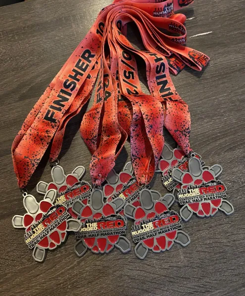 A pile of red and silver finisher medals for The Rugged Red Trail Half Marathon, with red ribbons that say "FINISHER".