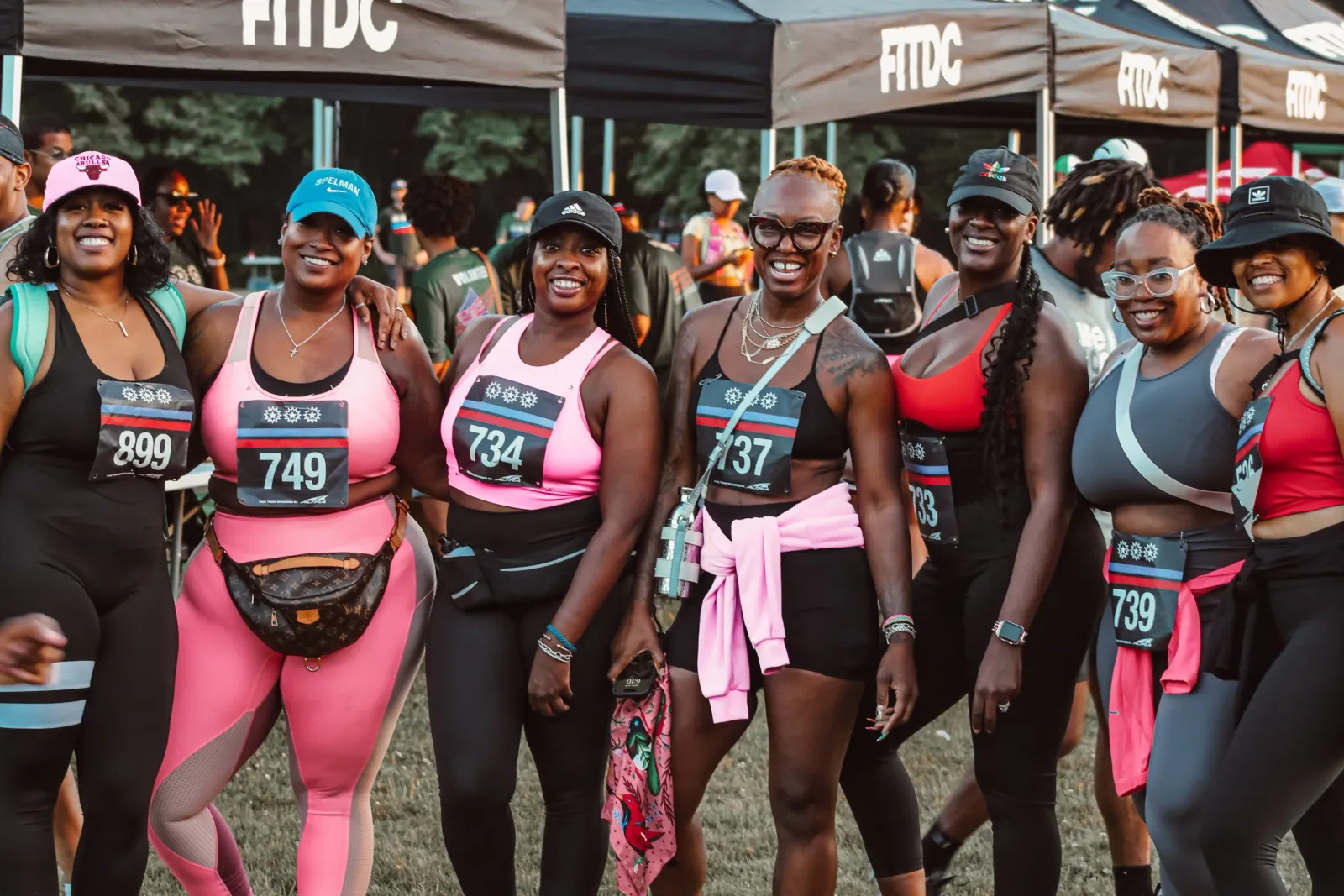 A group of diverse women smiling and posing for a photo at the start line of a race, wearing running attire and bib numbers.