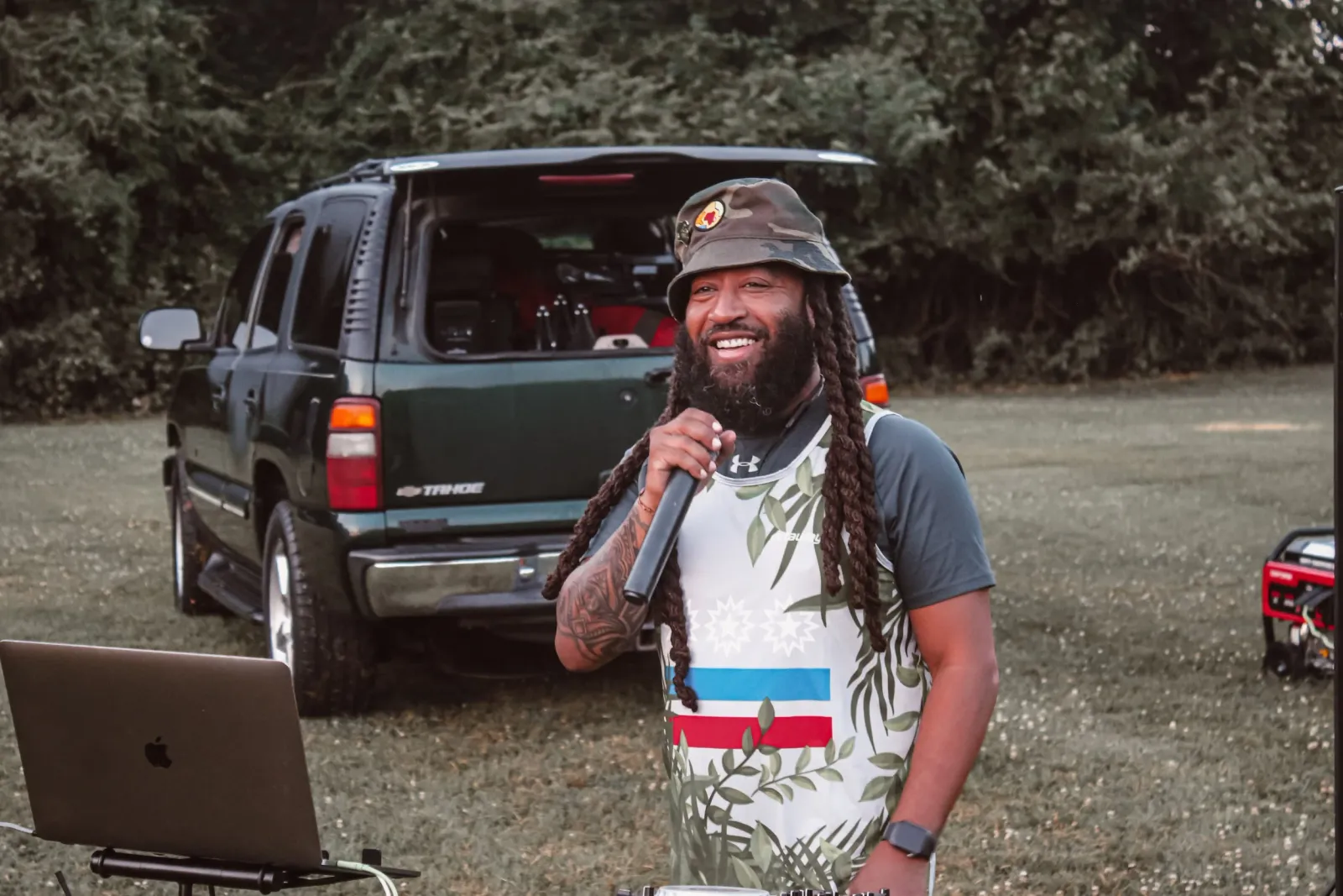 A smiling man with dreadlocks wearing a bucket hat and a colorful tank top holds a microphone, standing in front of an open SUV with a laptop and generator nearby.
